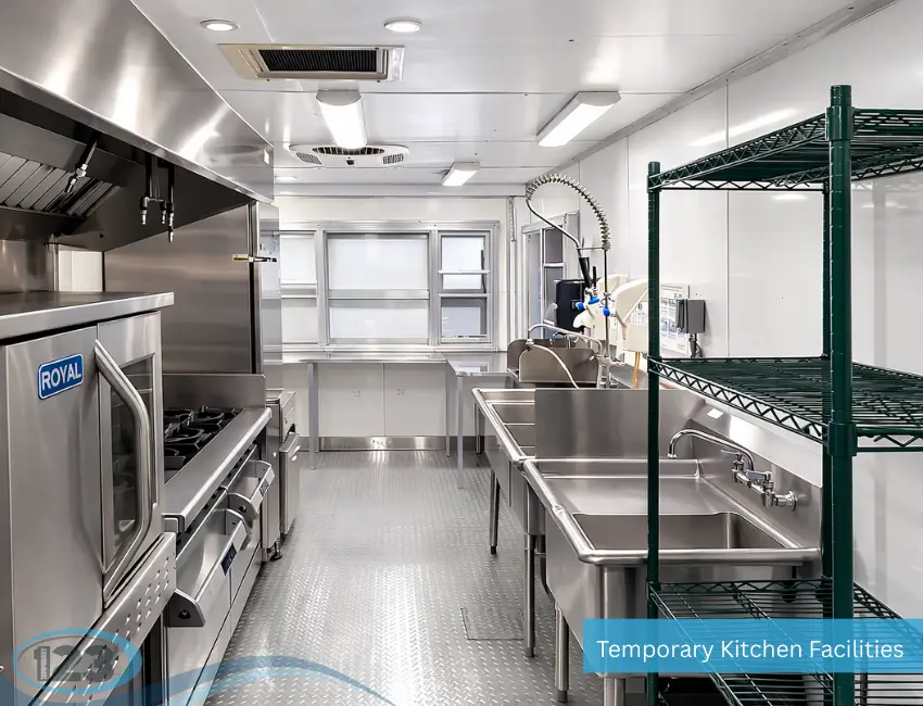An interior view of a bright, clean temporary kitchen facility featuring a commercial stove and oven on the left, multi-compartment stainless steel sinks, and green wire shelving on the right.