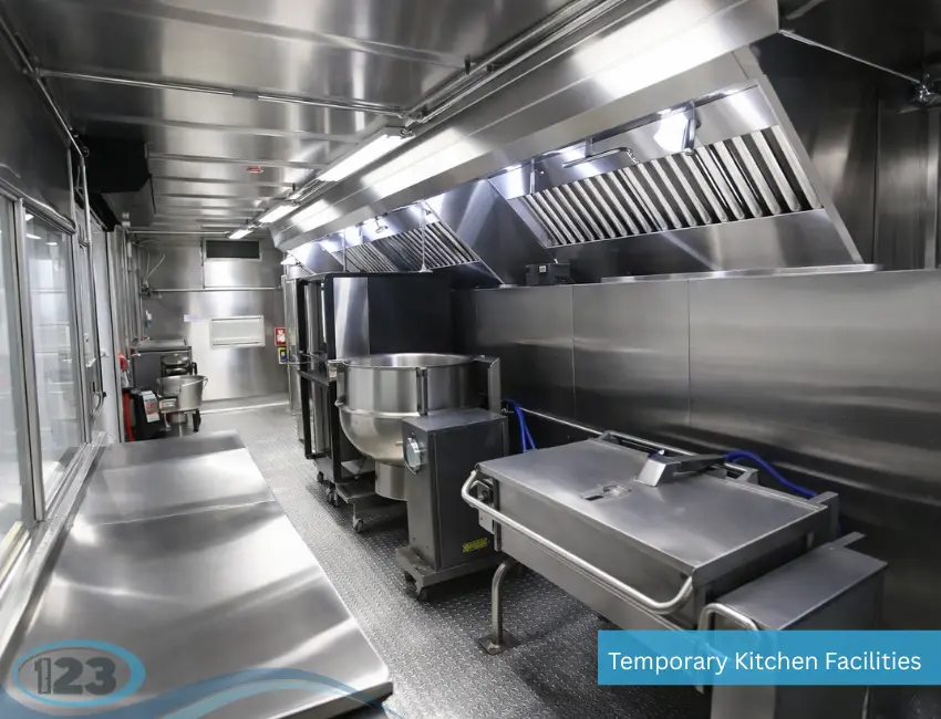 An interior view of a stainless steel temporary kitchen facility featuring heavy-duty cooking equipment, including a commercial tilt skillet, a large kettle, and overhead ventilation hoods.