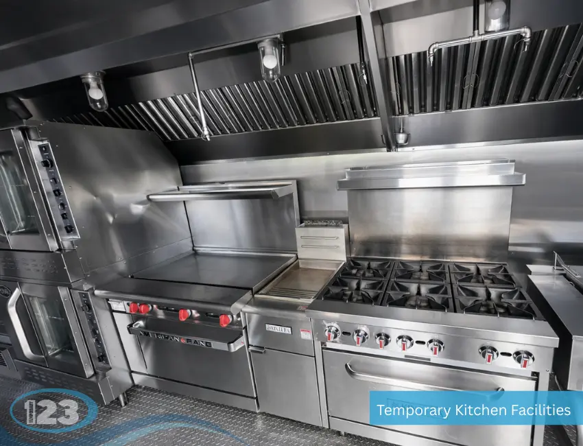 Stainless steel cooking equipment inside a temporary kitchen facility, featuring a commercial 6-burner stove, flat-top griddle, and overhead ventilation hoods.
