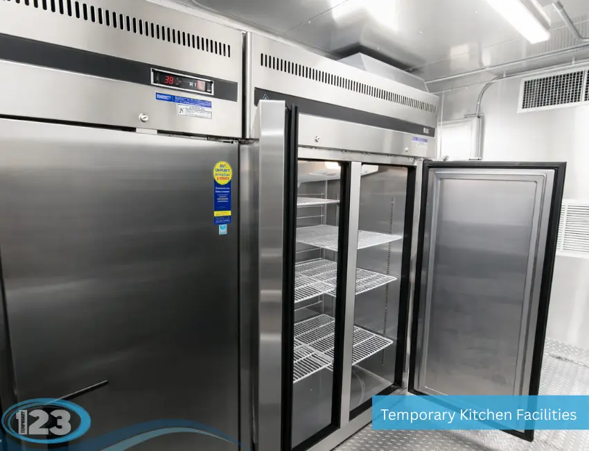 Commercial stainless steel refrigeration units inside a temporary kitchen facility, featuring a double-door reach-in refrigerator with its door open to show wire shelving.