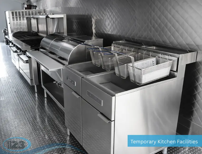 A row of commercial stainless steel kitchen equipment inside a temporary facility, featuring two deep fryers with wire baskets alongside a prep counter and cooking line.
