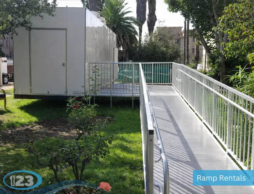 An exterior view of a long, metal wheelchair accessibility ramp with handrails leading up to the entrance of a temporary mobile trailer.